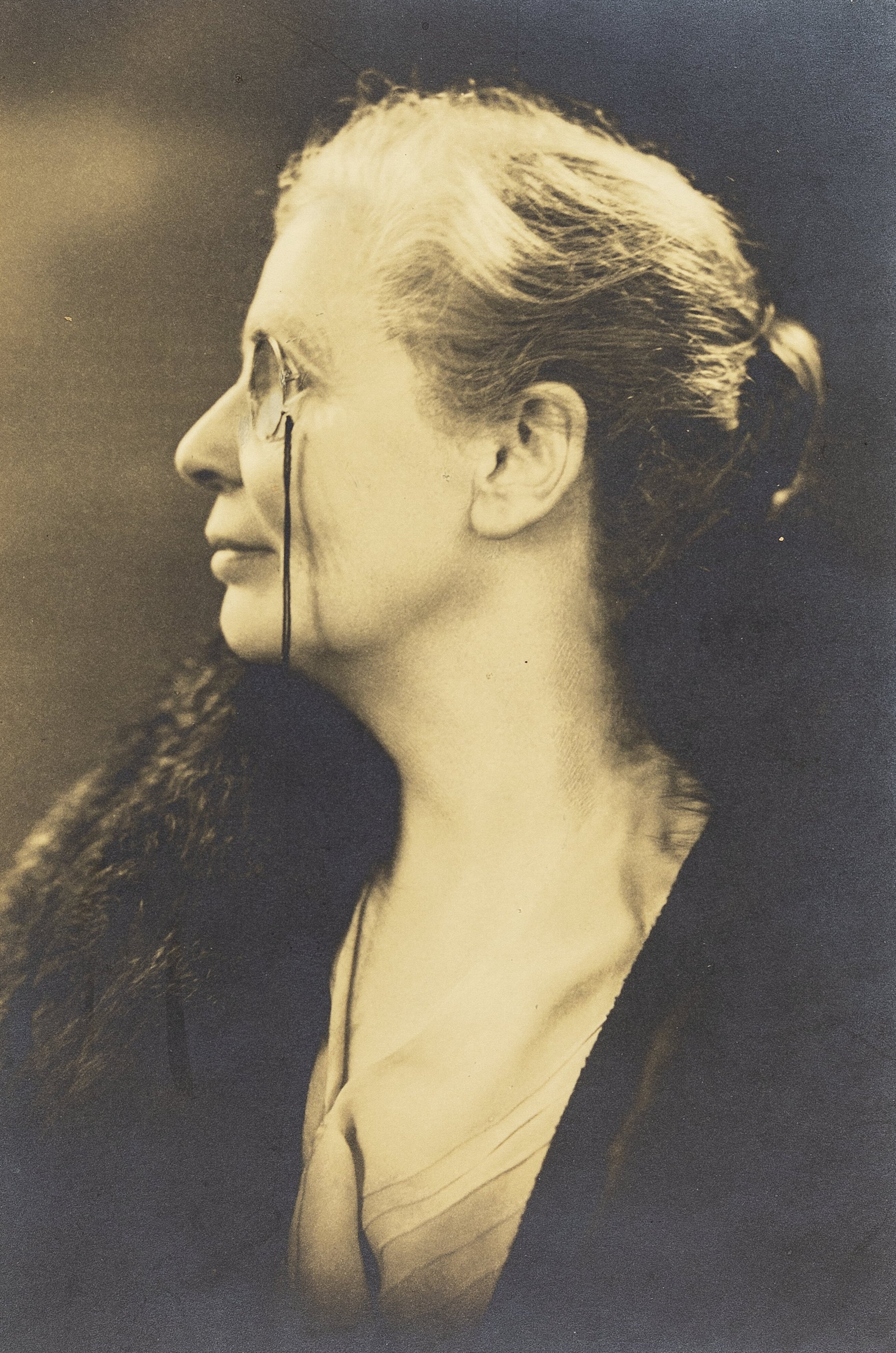 Monochrome studio portrait of Octavia Lewin, head and shoulders, left profile, wearing a monocle, mounted on card.